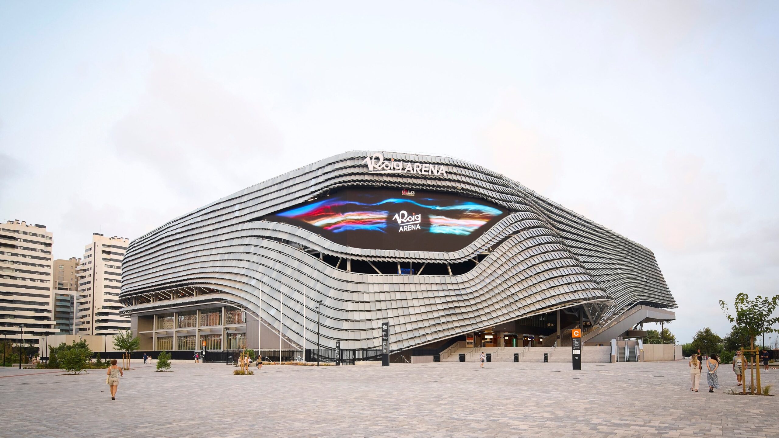 Roig Arena de València durante el día, edificio moderno y ondulado con fachada metálica y pantalla central, visto desde una plaza amplia con personas caminando alrededor.