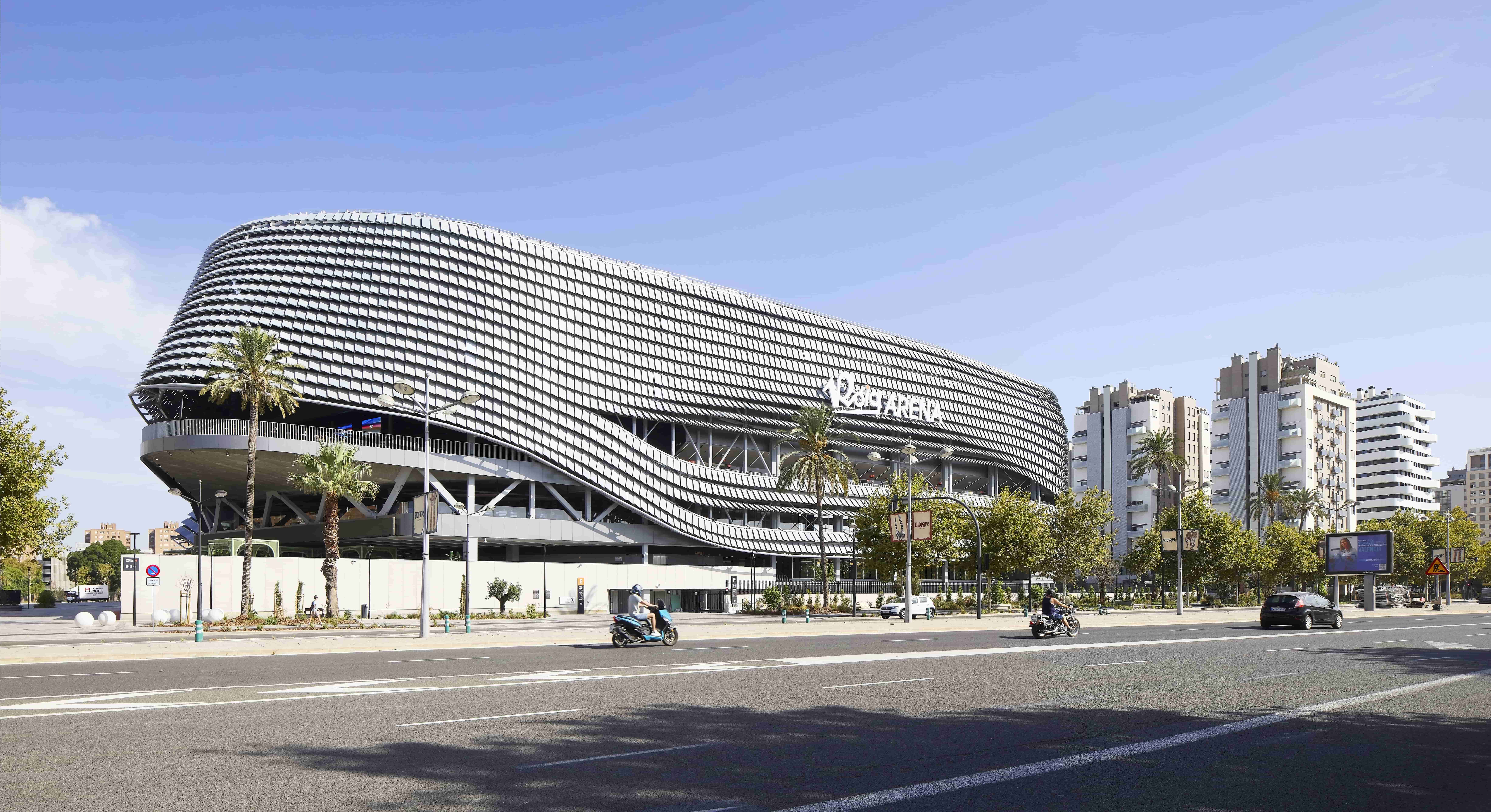 Vista exterior del Roig Arena de València de día, edificio moderno con fachada metálica ondulada junto a una avenida urbana con palmeras, tráfico y edificios residenciales alrededor.
