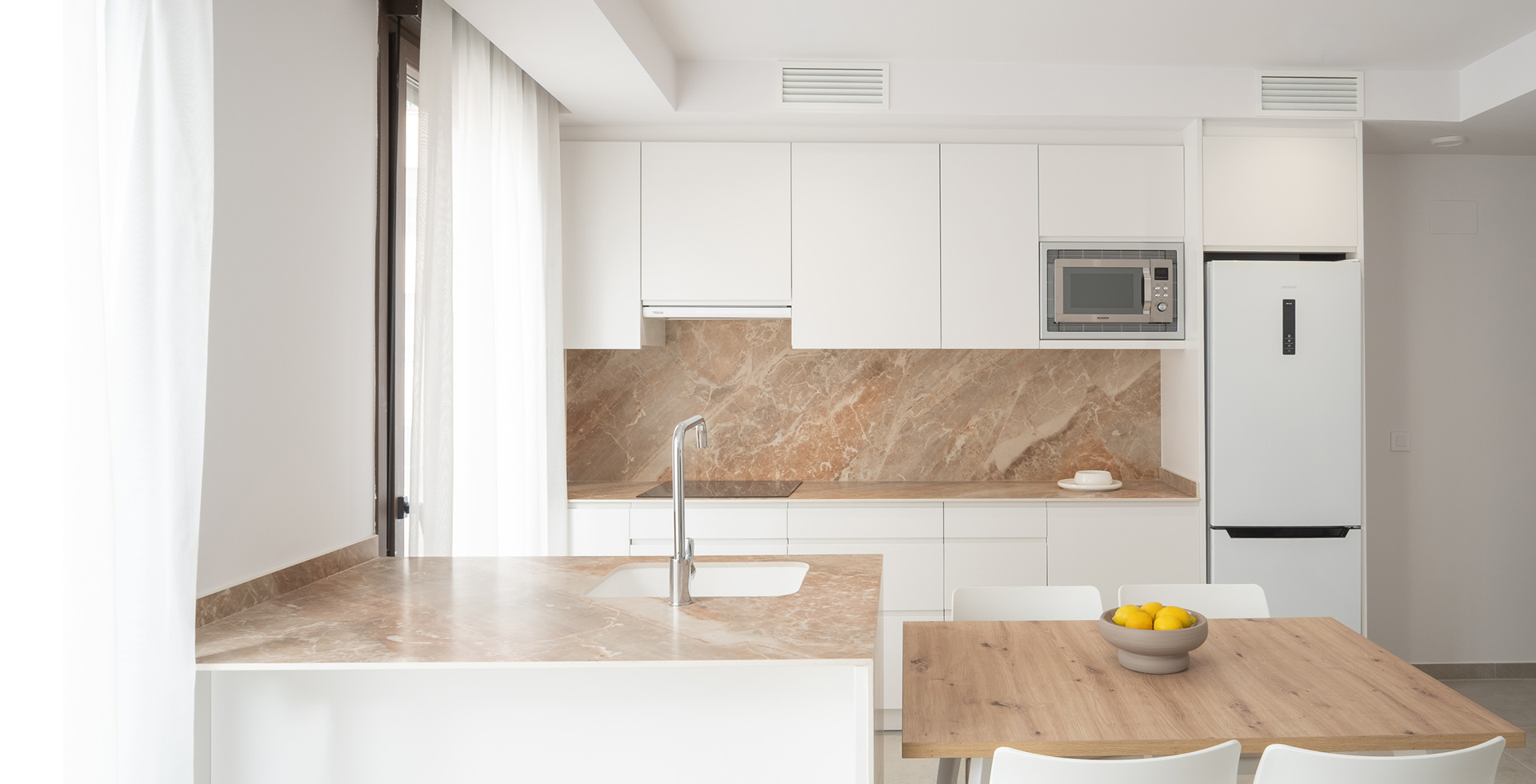 Interior de cocina de diseño contemporáneo con mobiliario blanco mate sin tiradores, salpicadero y encimera de mármol veteado en tonos tierra, península con grifería monomando cromada y mesa de comedor de madera de roble natural. La cocina esta situado en el interior del edificio Fola construido por OCN.
