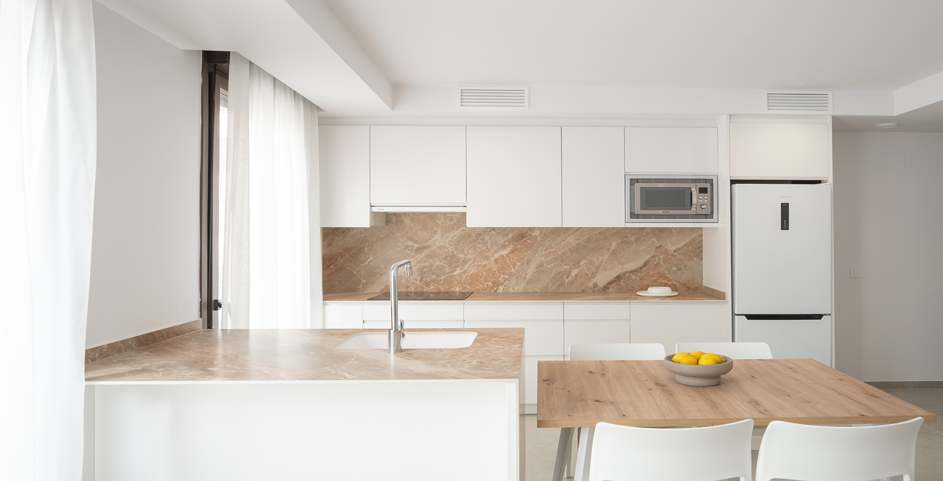 Interior de cocina de diseño contemporáneo con mobiliario blanco mate sin tiradores, salpicadero y encimera de mármol veteado en tonos tierra, península con grifería monomando cromada y mesa de comedor de madera de roble natural. La cocina esta situado en el interior del edificio Fola construido por OCN.
