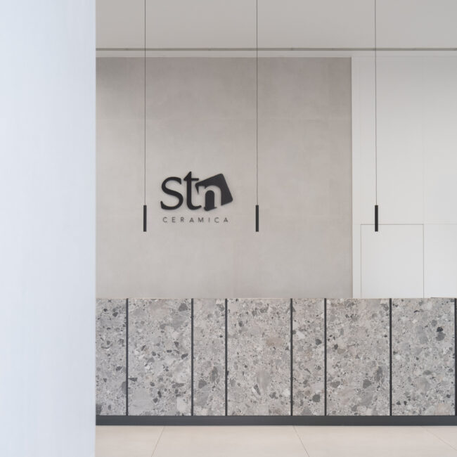Mostrador de recepción en STN Cerámica con revestimiento de terrazo técnico o piedra moteada, logotipo corporativo en relieve y luminarias suspendidas minimalistas.