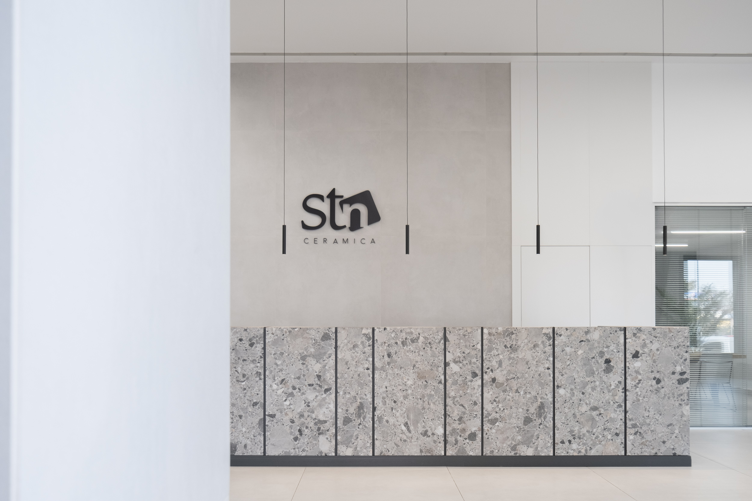 Mostrador de recepción en STN Cerámica con revestimiento de terrazo técnico o piedra moteada, logotipo corporativo en relieve y luminarias suspendidas minimalistas.
