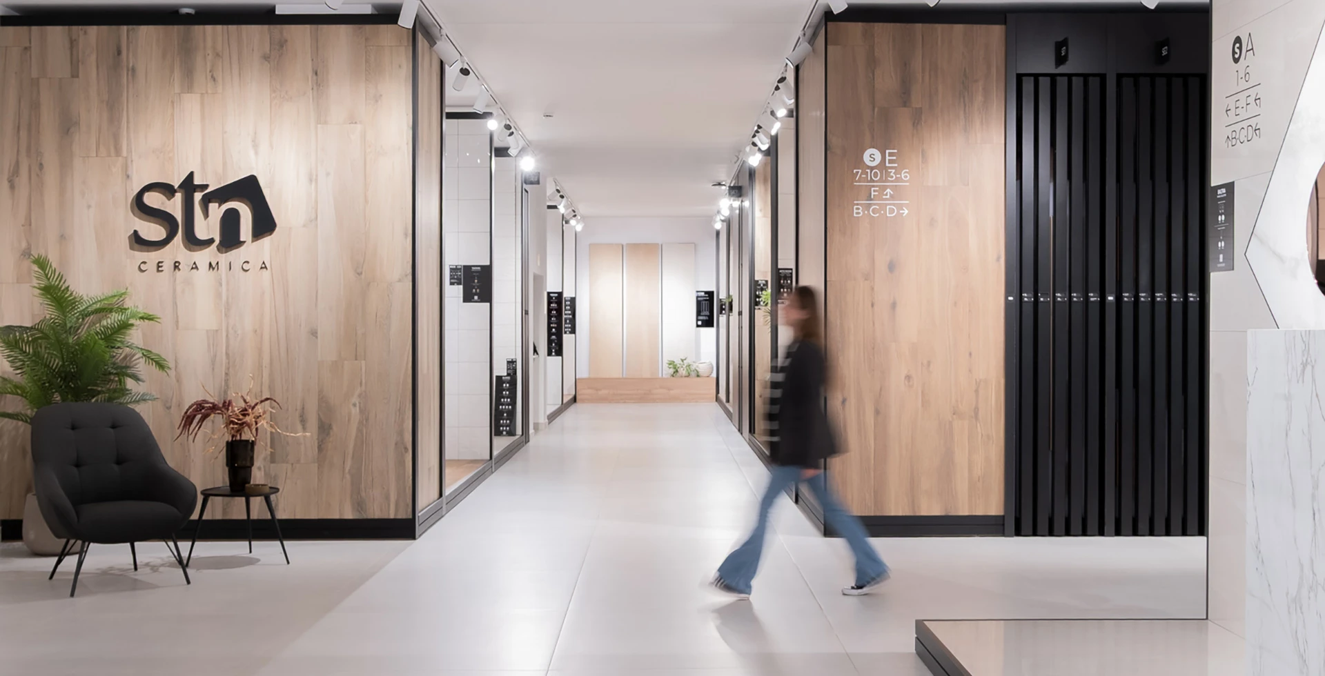 : Showroom de azulejos con diseño moderno: pasillo con paneles efecto madera, iluminación técnica de carril, sillón de diseño y logotipo de STN Cerámica.
