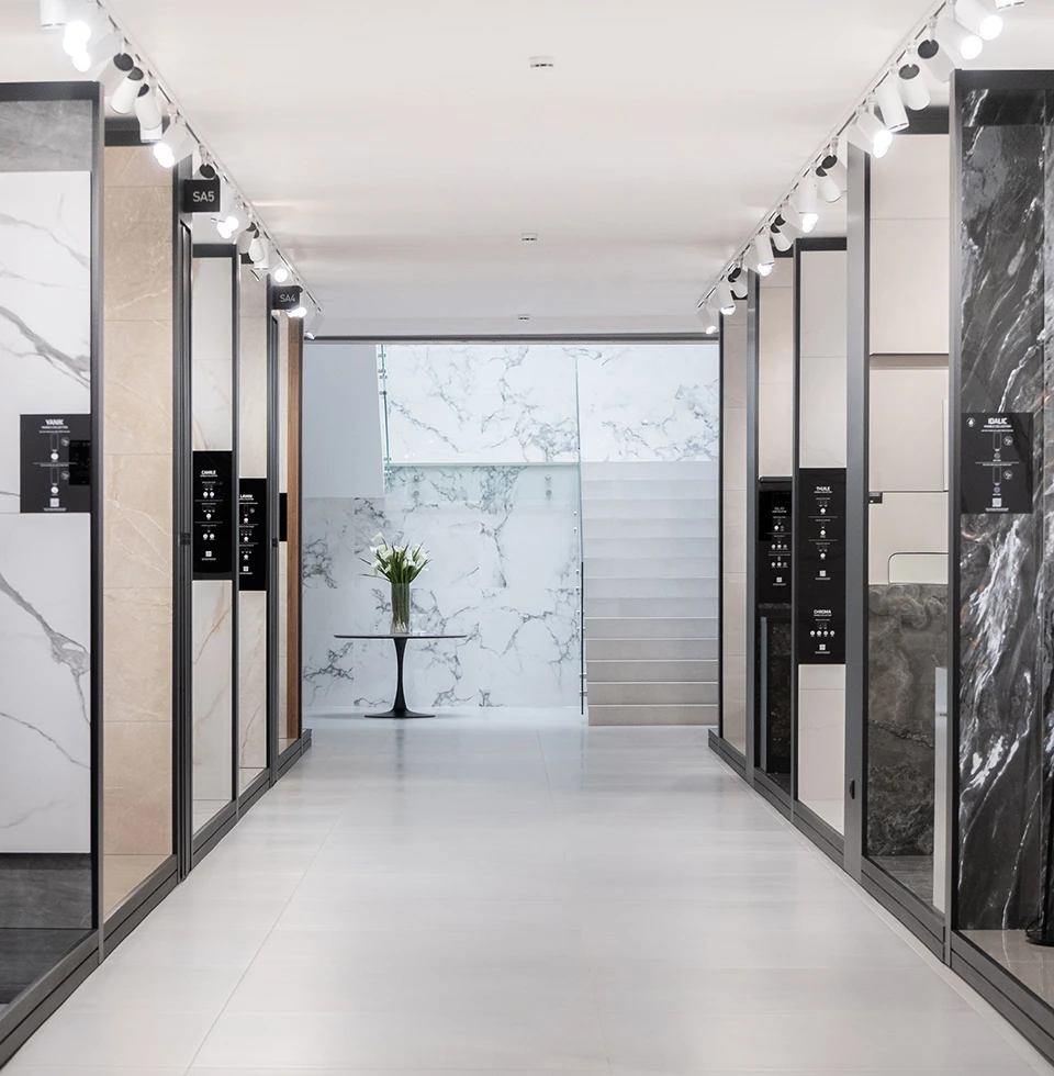 Showroom cerámico con paneles expositores de mármol de gran formato, iluminación técnica de focos en carril y escalera integrada en el diseño del espacio.