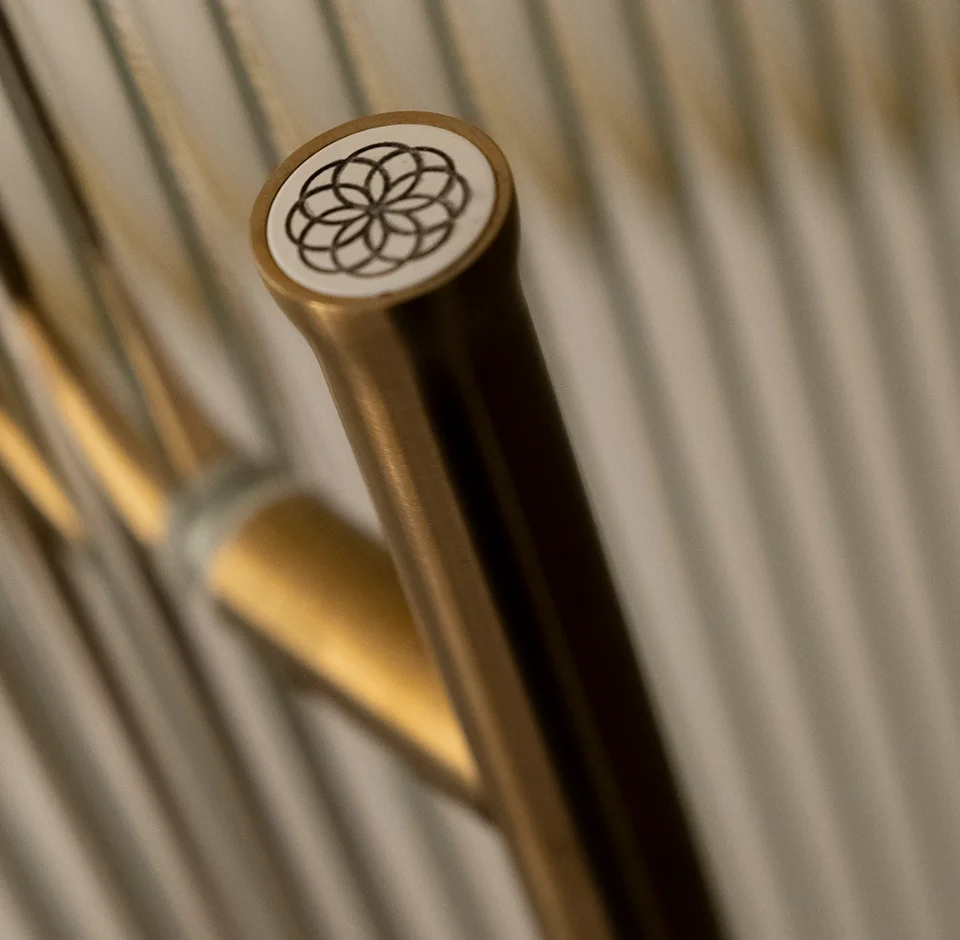 Primer plano de un elegante pomo cilíndrico de metal con acabado en latón cepillado o bronce. En la parte superior plana de color blanco, destaca un intrincado grabado negro con un motivo geométrico de círculos entrelazados (flor de la vida). El fondo muestra una superficie estriada con líneas verticales metálicas desenfocadas, resaltando la precisión técnica y el diseño de autor presente en la constructora de hoteles de lujo OCN en Valencia